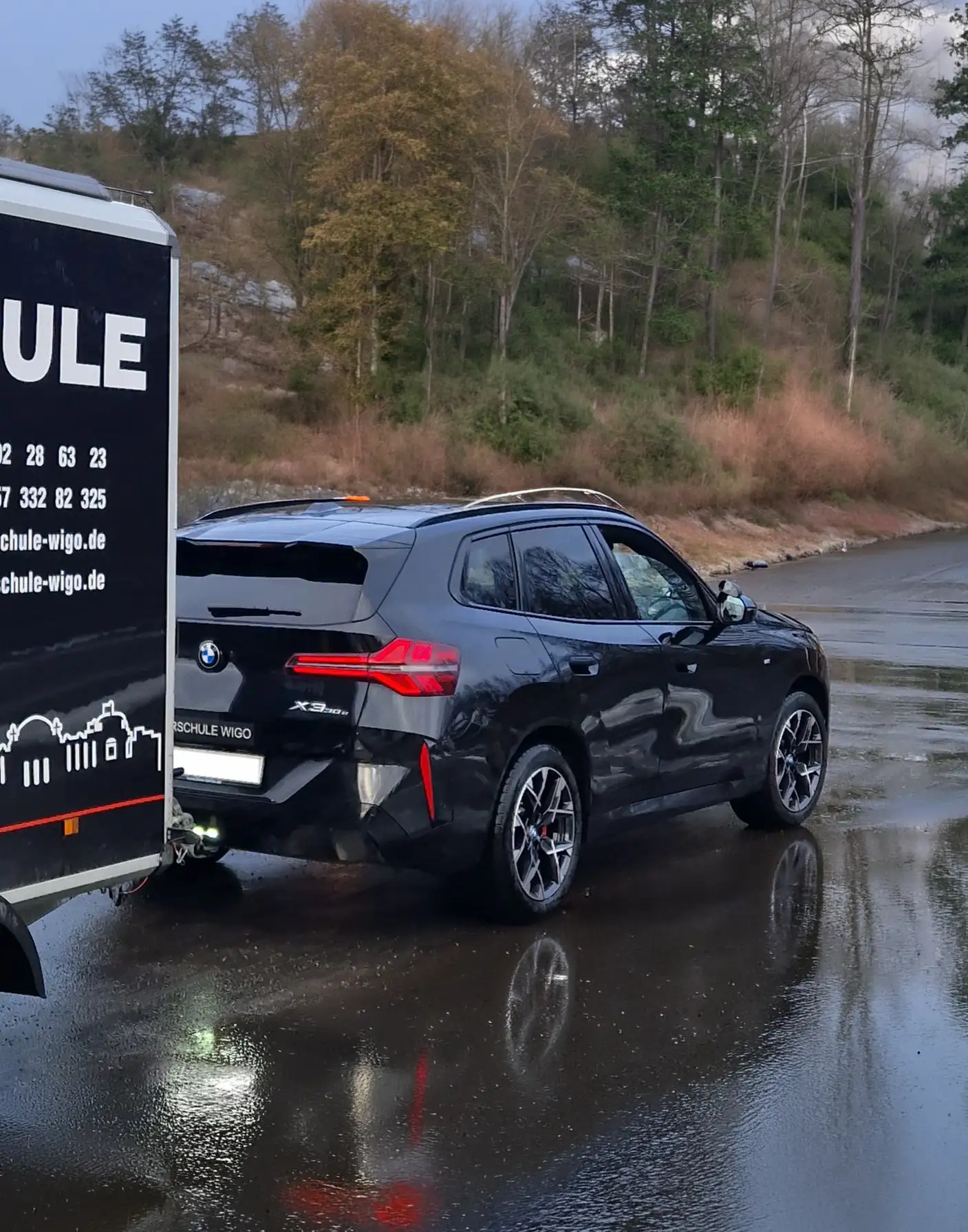 Schwarzer BMW X3 mit Anhänger der Fahrschule WiGo auf einer nassen Straße